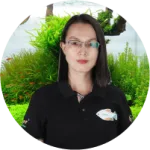A woman wearing a black polo shirt with a fish logo, standing in front of lush green plants and a fish tank.