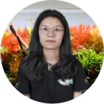 A woman with long black hair wearing a black polo shirt stands in front of vibrant aquarium plants with red and green foliage