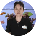 A woman in a black polo shirt with a fish logo stands in front of a vibrant aquarium display featuring colorful coral and fis