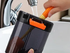 A hand using an orange brush to clean a black and orange hygger aquarium filter under running water in a bathroom sink.